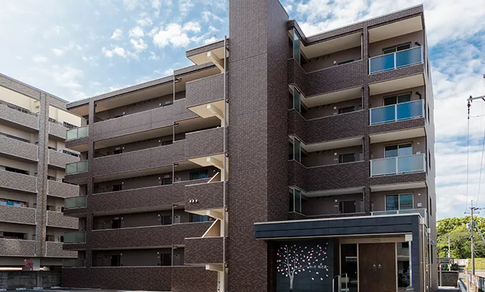 青空と青々とした木々に包まれたブラウンタイルのモダンな6階建てマンション外観全景
